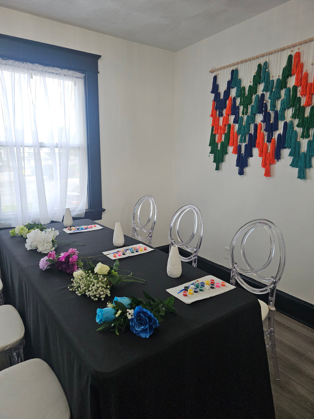 A styled event table setup in a room with white walls, featuring a black draped tablecloth, clear acrylic chairs, floral centerpieces, and a colorful woven wall hanging on the wall.